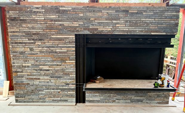 Partially constructed exterior fireplace surround clad in stacked thin stone veneer, with a large black firebox opening and a stone hearth. The image is taken on a building site, with exposed framing and construction tools visible around the installation.