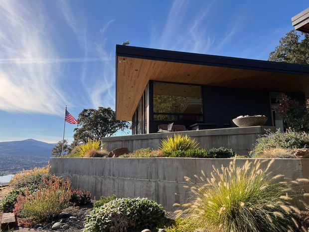 Exterior view of a contemporary single-storey home on a sloped, terraced site. The house has a flat roof with deep overhangs, dark horizontal cladding, extensive glazing, and a warm timber-lined soffit. Layered concrete retaining walls and drought-tolerant ornamental planting form the landscaped foreground, with a distant mountainous landscape visible beyond.