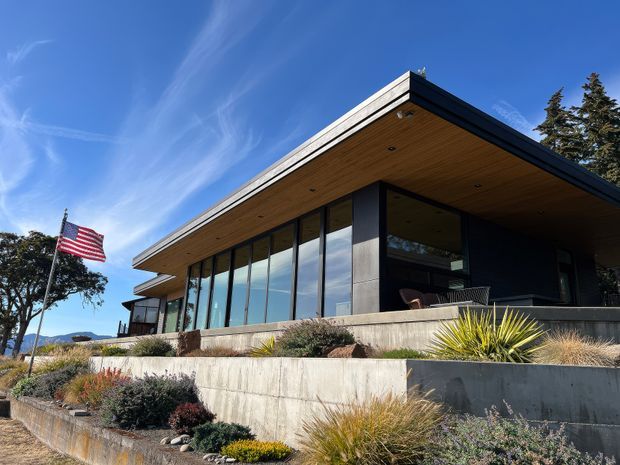 Modern single-storey residence with a broad flat roof, deep cantilevered eaves, dark exterior walls, extensive floor-to-ceiling glazing, and a warm timber-lined soffit. The home sits on a terraced, sloping landscaped site with concrete retaining walls and drought-tolerant planting. An American flag is visible beside the house.