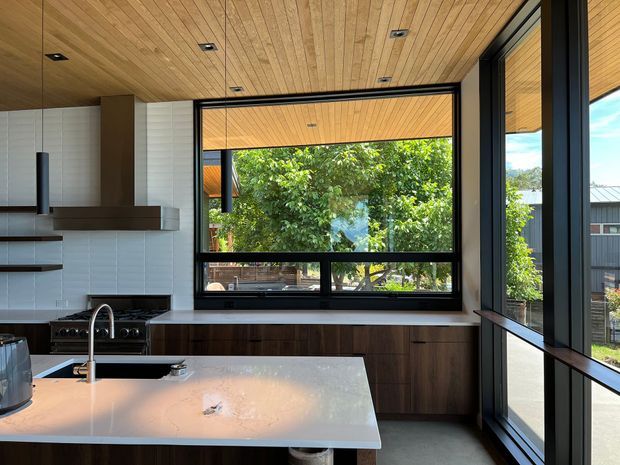Contemporary kitchen with a large central island, dark timber cabinetry, white stone benchtops, integrated cooktop and sink, white tiled splashback, and expansive black-framed glazing. The ceiling is lined with warm timber boards, and the windows overlook leafy outdoor surroundings.