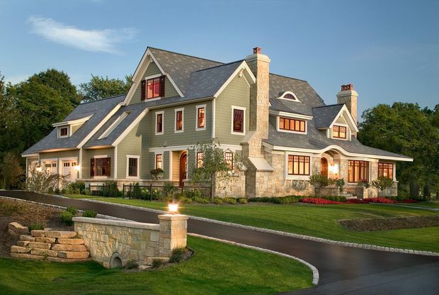 Exterior view of a large, multi-wing country-style residence with steep intersecting gabled roofs, grey shingle cladding, stone chimneys and lower stone wall sections. The home has many divided-light windows with warm interior lighting, landscaped lawns, curved paved driveways, and a low stone bridge or culvert in the foreground.