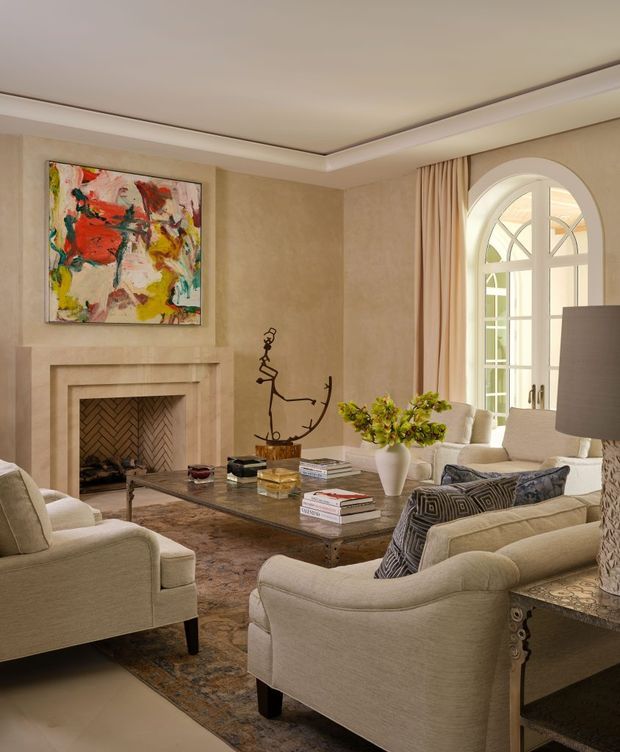 Elegant, traditionally styled living room with warm beige plaster walls, layered crown moulding and recessed ceiling detailing. The room features a substantial stone or plaster fireplace with a herringbone brick-lined firebox, an abstract colourful artwork above the mantel, several cream upholstered armchairs and sofas, a dark glass-topped coffee table, patterned rug, sculptural wall artwork, and tall arched multi-pane windows with full-height curtains. The overall atmosphere is refined, warm, and inviting.