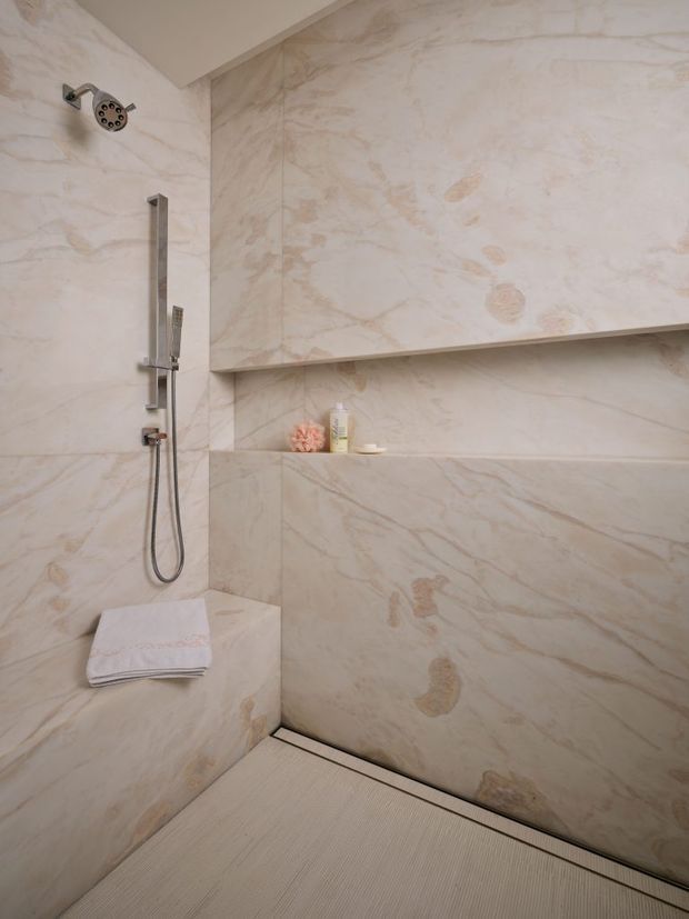 A luxurious walk-in shower clad entirely in pale cream and beige veined natural stone, with a rain showerhead, handheld shower on a vertical rail, recessed wall niches, a built-in corner bench, and a linear floor drain. Chrome fixtures and neatly folded towels add functional detail.