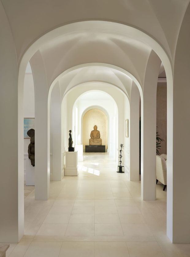 A serene, grand interior hallway or gallery with a sequence of white rounded arches and vaulted ceilings. The long corridor is finished in pale tones with large light-coloured floor tiles, sculptural artwork, a Buddha statue at the far end, and strong natural light creating a calm, gallery-like atmosphere.