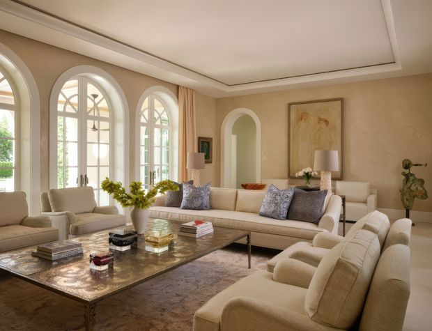 Elegant, spacious living room with a warm neutral palette, arched floor-to-ceiling windows and matching arched wall niche, layered cream seating, patterned cushions, a large stone or marble coffee table, soft rug, artwork, and refined decorative styling. The recessed tray ceiling and classical arches create a polished, traditional luxury character.