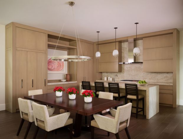 A warm, open-plan kitchen and dining area with extensive light timber cabinetry, a dark rectangular dining table surrounded by upholstered chairs, and a kitchen island with bar seating. Pendant lights hang above both the dining table and island. The kitchen includes a patterned stone backsplash, integrated appliances, open shelving, and dark timber flooring.