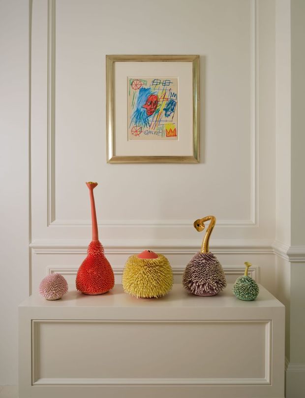 A refined interior vignette featuring a white paneled wall and built-in console, topped with a playful collection of brightly coloured textured sculptural vessels. A framed childlike artwork hangs centrally above the display. The composition combines classic architectural mouldings with whimsical contemporary objects.