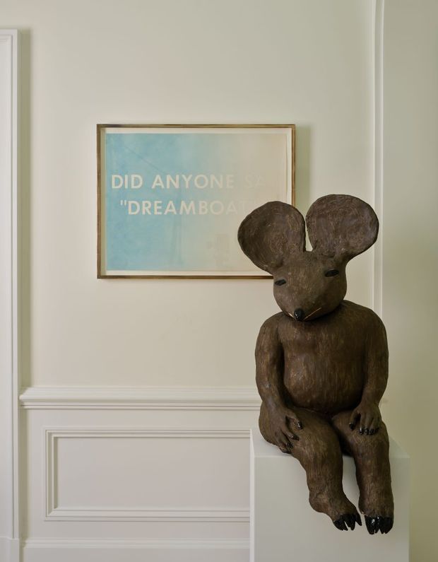 A quiet, refined interior vignette featuring a large dark wood-toned mouse sculpture seated on a white plinth against a pale beige wall. The wall has traditional raised-panel wainscoting and crisp white trim, with a framed blue artwork above the sculpture. The composition is softly lit and understated, with a whimsical sculptural accent.
