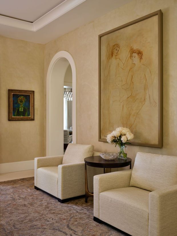 A refined sitting area with two cream upholstered armchairs arranged around a dark round side table. The room features warm beige textured wall finishes, white decorative ceiling trim, a patterned neutral rug, an arched doorway, framed artwork, and a vase of flowers. The overall atmosphere is elegant, warm, and softly traditional.