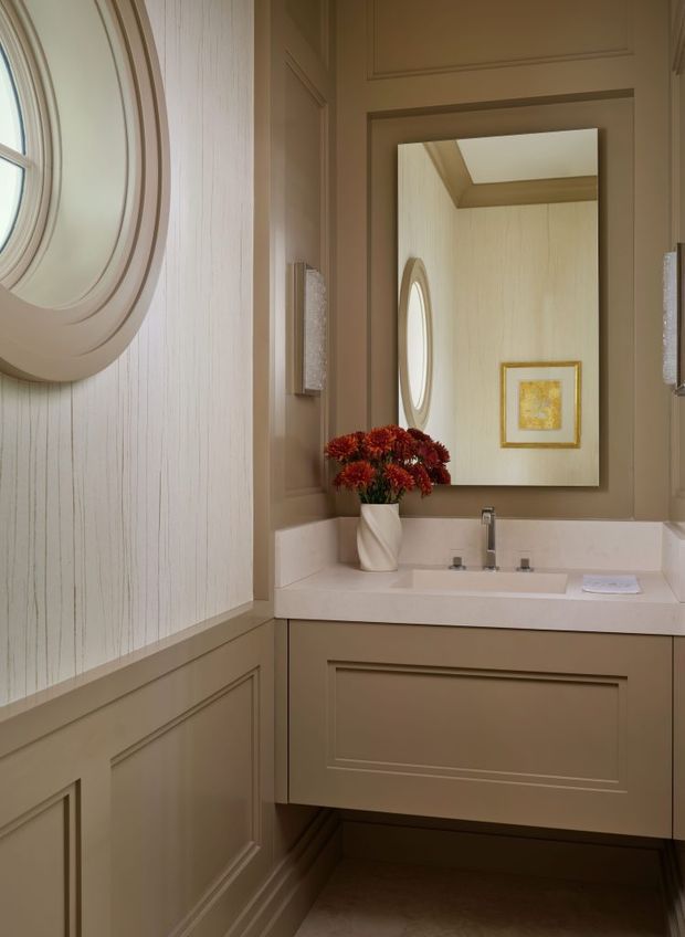 A compact, elegant powder room with taupe-beige raised-panel cabinetry, a white integrated vanity basin and polished metal tapware. The walls feature vertical textured wallpaper above detailed wainscoting and panel mouldings. A large rectangular mirror reflects an oval window, framed artwork and the vanity, while wall sconces and a vase of flowers add decorative detail. The room has a warm, refined traditional aesthetic.