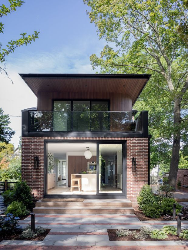 Front exterior view of a compact two-storey residential extension or house, featuring a dark low-pitched roof with a broad overhang, warm timber soffit and upper-level cladding, a black-framed glazed balcony, and a red brick lower facade. Large sliding glass doors reveal a bright kitchen inside. Timber steps lead to a landscaped paved garden path with established trees and shrubs.