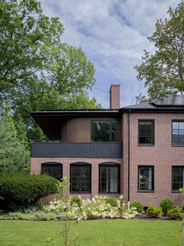 Exterior view of a multi-storey brick house set within a landscaped garden. The facade combines red brickwork with dark-framed windows, a projecting upper-level balcony or terrace with dark vertical railing, dark roof edges, and a prominent brick chimney. Mature trees and planted beds surround the building.