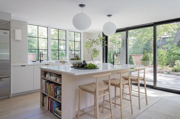Bright, airy contemporary kitchen with a large white island and integrated seating for four. The room features pale cabinetry, light timber flooring, white benchtops, black-framed steel-look windows and extensive sliding glazing opening to a leafy garden. Open shelving at the island displays books and kitchenware, while two large spherical pendant lights hang above the island.