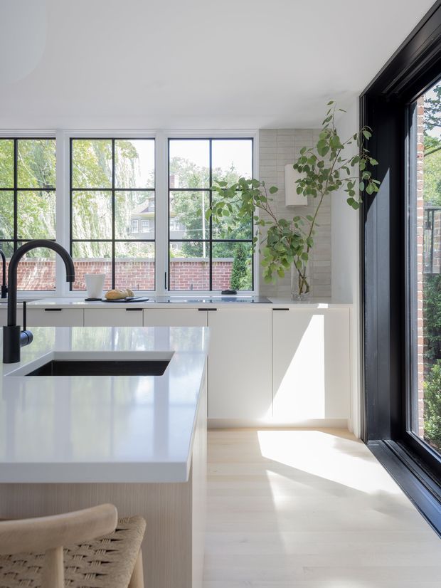 Bright contemporary kitchen with a large white island and integrated dark sink, white flat-panel cabinetry, pale timber flooring, and a light stone or engineered-stone benchtop. Black-framed industrial-style grid windows and full-height glazing bring in abundant daylight, while a light tiled feature wall and indoor tree add texture and natural character.