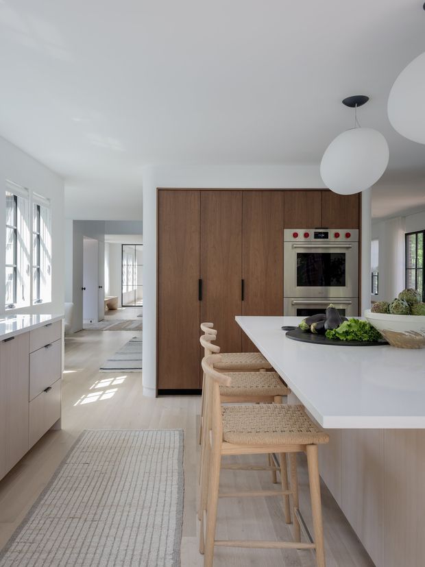 Bright contemporary kitchen with a large white island and integrated seating, warm wood full-height cabinetry, built-in stainless-steel oven, pale flooring, and abundant natural light from tall windows. The open-plan space extends toward a hallway and adjoining areas, with sculptural white pendant lights overhead.