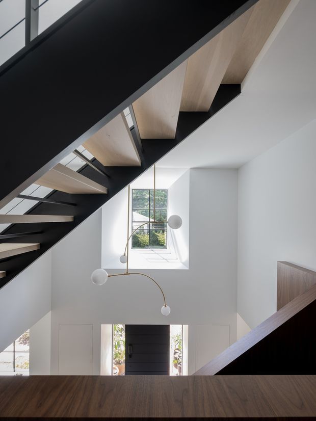 A bright, minimalist stairway viewed from an upper landing, featuring a dramatic black-painted stair structure with pale timber treads, white walls, tall windows, and a sculptural hanging globe light. A dark timber landing or handrail occupies the foreground, while the entry door and glazing are visible below.