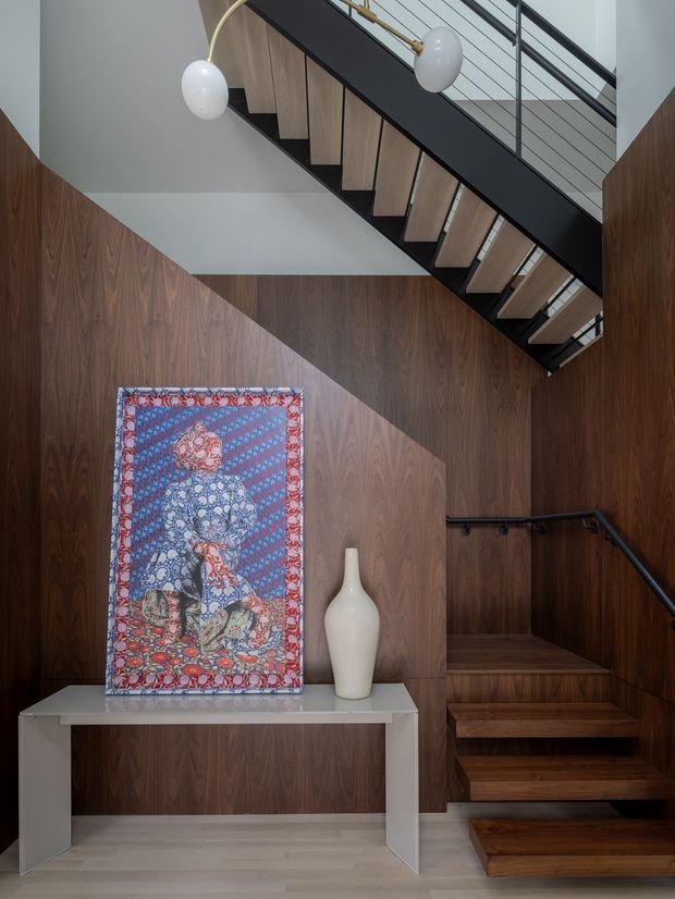 A refined residential stairway with dark timber wall panelling, matching timber stair treads and a black metal balustrade. A slim pale console table displays a colourful textile artwork and white ceramic vase. Sculptural globe pendant lights hang above, while natural light creates a geometric shadow across the wall.