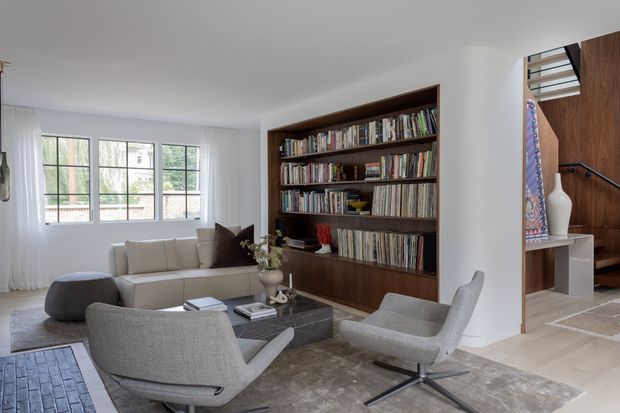 A bright, refined living room with large gridded windows dressed in sheer curtains, a light upholstered sofa, sculptural lounge chairs, a low dark coffee table, and a large area rug. A full-height dark timber built-in bookshelf forms a strong feature wall, while an adjacent open stair with dark timber wall panelling and a black handrail is visible to the right. The space has a calm contemporary feel with natural daylight and warm wood accents.