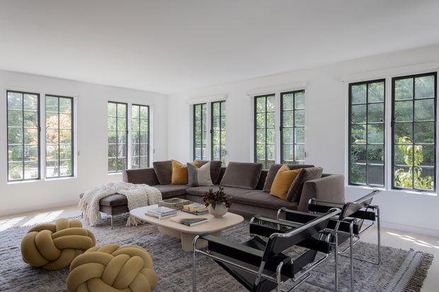 Bright, spacious contemporary living room with white walls, high ceilings, extensive black-framed multi-pane windows, and views of greenery outside. A large taupe sectional sofa surrounds a rounded light coffee table, with sculptural mustard lounge chairs, black leather sling chairs, layered cushions, and a textured grey rug. The room has a refined, comfortable, modern aesthetic with strong natural light.