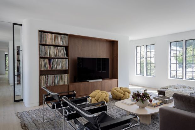 Bright contemporary living room with a built-in dark timber entertainment wall, open vinyl-record shelving and television, large multi-pane windows, sculptural lounge chairs, a sectional sofa, and a layered neutral rug and coffee table. The room has a refined, curated feel with strong natural light and a spacious layout.