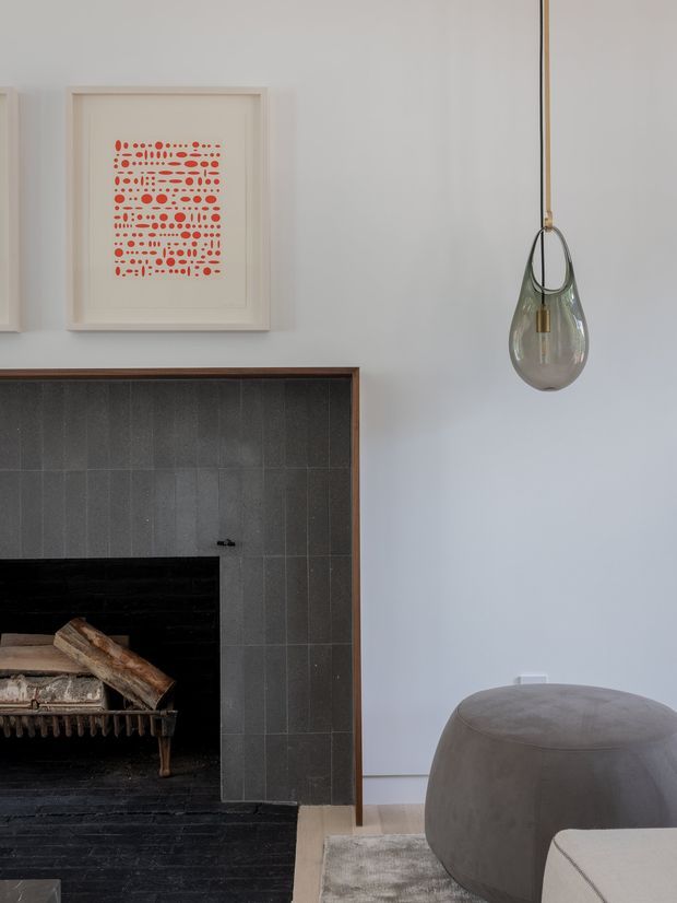 A refined living room vignette featuring a dark tiled fireplace surround with a wood-burning firebox, pale walls, framed abstract artwork, a sculptural pendant light, and a large rounded upholstered ottoman. The palette is restrained and neutral with charcoal, white, taupe, and muted timber accents.