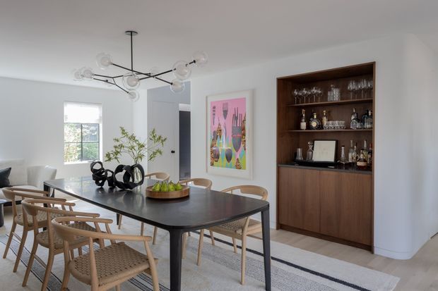 A bright, refined dining room with a large dark rectangular dining table surrounded by woven-seat timber chairs. The room features white walls, pale flooring, a sculptural black-and-clear glass chandelier, colourful framed artwork, and a substantial built-in dark timber drinks cabinet with open shelving displaying glassware and bottles. A window brings in natural light, and a doorway connects to an adjacent area.