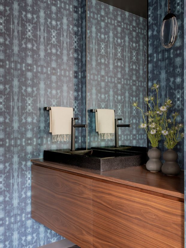 A richly styled bathroom vanity featuring a floating dark timber cabinet, a black rectangular vessel basin with matching dark fixtures, and a large mirrored wall. The walls are covered in a blue-grey patterned finish, with cream hand towels and sculptural dark vases containing delicate dried floral stems. The mood is dark, refined, and luxurious.