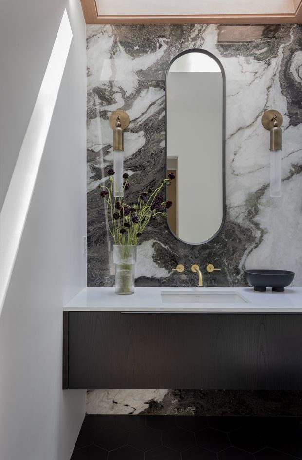 A refined contemporary bathroom vanity featuring a dramatic grey-and-white veined stone wall, a dark floating timber vanity with a white benchtop and integrated basin, brushed brass wall-mounted tapware and sconces, and a tall arched mirror. Black geometric floor tiles and warm timber ceiling trim add contrast.