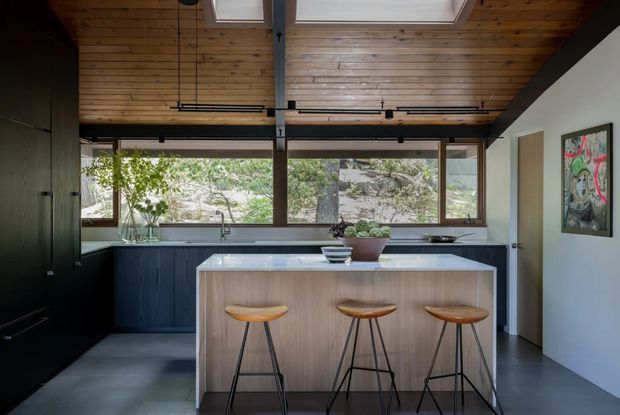 A contemporary kitchen with a large central island and three bar stools, dark fixed cabinetry, pale timber island joinery, and a dark polished-looking floor. Expansive horizontal windows overlook rocky, wooded terrain, while exposed timber ceiling boards, black structural framing, and two skylights add architectural character. The space is clean and understated with a warm timber and charcoal palette.