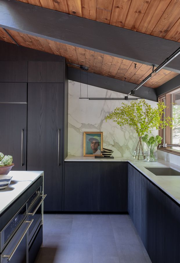 A sophisticated contemporary kitchen with dark charcoal-black timber cabinetry, integrated tall storage, pale stone benchtops and a marble-look stone feature wall. The room features a warm timber-lined ceiling with substantial dark exposed beams, slim black track and pendant lighting, a large window, and restrained decorative styling with greenery and artwork. Grey floor tiles and dark joinery create a moody yet refined atmosphere.
