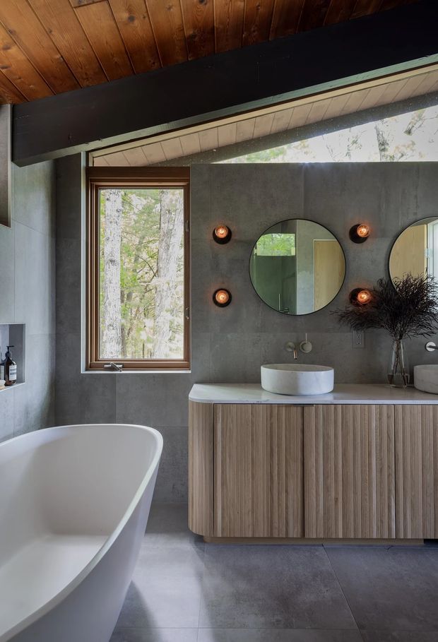 A sophisticated bathroom with a freestanding white bathtub, a floating fluted-wood vanity with a pale stone benchtop, round vessel basin, twin circular mirrors, and warm globe wall lights. The room features grey concrete-look wall and floor finishes, dark structural beams, a timber-lined pitched ceiling, and a timber-framed window overlooking trees.