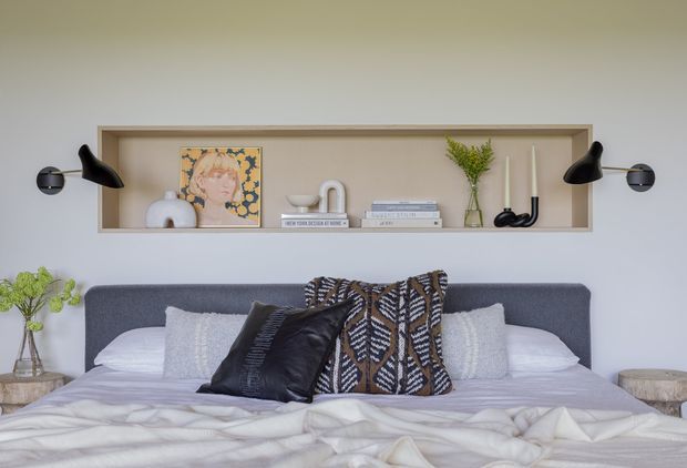 A styled bedroom with a neatly made bed, layered white and grey bedding, textured decorative cushions, and a dark woven throw. A recessed wall shelf behind the bed displays artwork, ceramics, books, and a vase with greenery. Black wall-mounted swing-arm lamps flank the shelf, creating a refined contemporary composition in soft neutral tones.