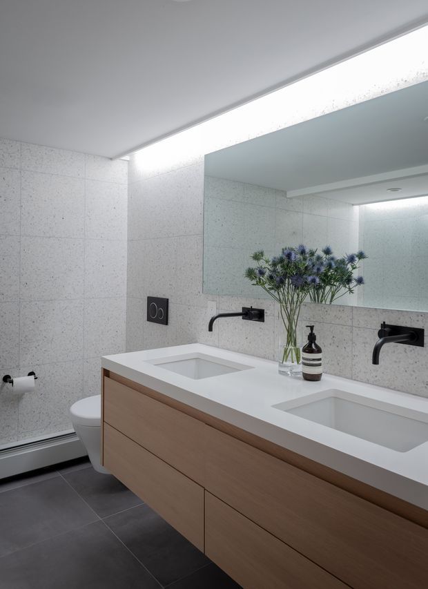 Contemporary bathroom with a double floating timber vanity, white integrated basins and benchtop, matte black wall-mounted tapware, large frameless mirror, pale speckled wall tiles, and dark floor tiles. A toilet and heated towel rail are visible at the side, with a vase of flowers and soap dispenser on the vanity.