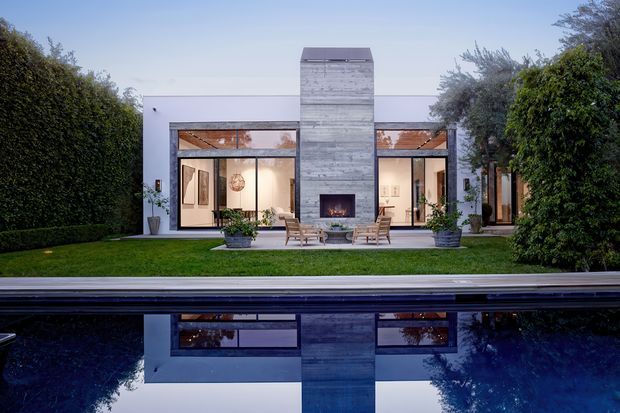 A low, contemporary residence viewed across a reflecting swimming pool. The white rectilinear facade has extensive glazed openings, a prominent central grey stone-clad chimney or fireplace tower, and a sheltered outdoor seating area facing the pool. Manicured hedges and trees frame the building.