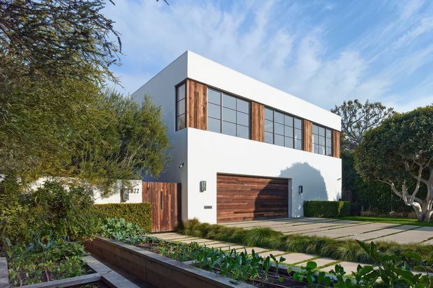 A modern two-storey residential house with a crisp white rendered cubic facade, extensive upper-level grid windows, vertical timber accents, and a timber-clad garage door. The property is viewed from a landscaped front garden with hedges, raised planting beds, and a broad paved driveway. The composition is contemporary and minimalist, with strong geometric lines and warm wood contrasting against the white exterior.