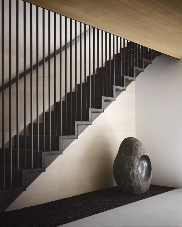 A dramatic modern stairway with dark metal vertical balusters and zigzag-edged stair stringer beneath a warm timber-lined ceiling. The staircase runs diagonally across a pale textured wall, with a sculptural stone object displayed on a dark gravel-like floor strip below.