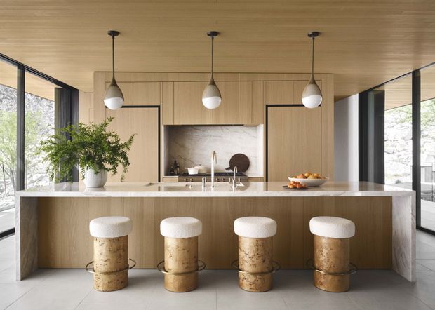 A refined contemporary kitchen with a large waterfall-edge island and four upholstered stools. The space features warm timber cabinetry and ceiling lining, a pale stone benchtop, integrated stainless-steel sink and tapware, a stone splashback, black-framed floor-to-ceiling glazing, and three sculptural pendant lights. The open glazing brings in abundant natural light and views of trees and stonework outside.