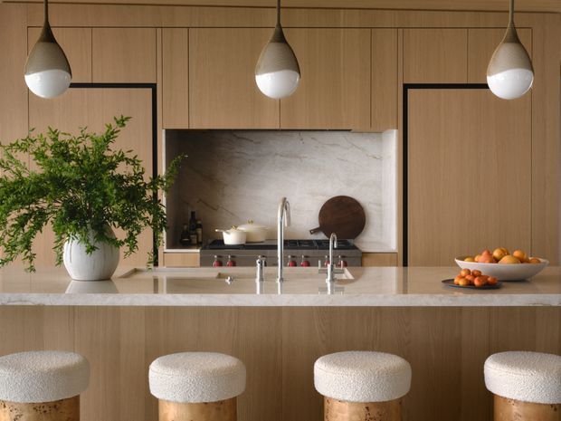 Warm contemporary kitchen featuring full-height light timber cabinetry, a large pale stone island bench with integrated sink and tapware, a matching stone backsplash, and four upholstered timber bar stools. Three sculptural pendant lights hang above the island, while greenery and serving bowls add natural accents.