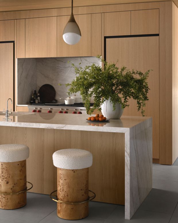 A refined contemporary kitchen featuring warm vertical timber cabinetry, a large stone-clad island with waterfall edge, integrated cooktop and sink, and a light veined stone splashback. Sculptural pendant lighting, rounded upholstered stools with timber bases, and a large leafy arrangement add softness and warmth.