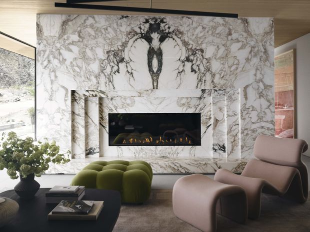Luxurious contemporary living room featuring a dramatic bookmatched marble fireplace wall with integrated linear fire, sculptural lounge chairs, a rounded green sofa, dark coffee table, and warm timber ceiling and floor finishes. Large glazing brings in a bright natural landscape view.