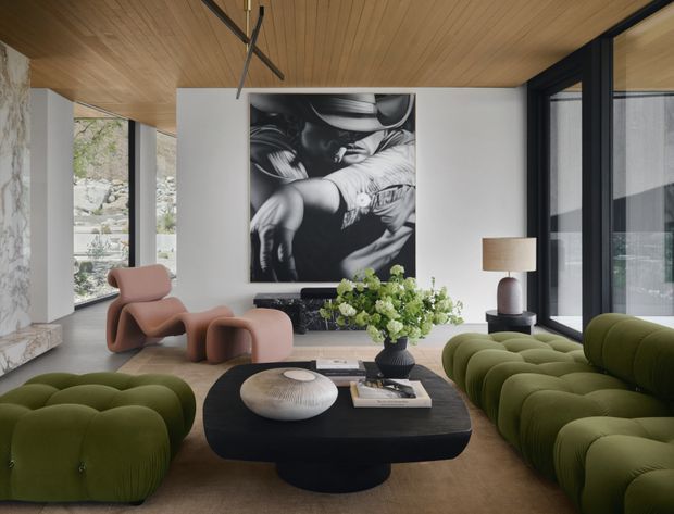 A sophisticated contemporary living room with floor-to-ceiling glazing, a warm timber-lined ceiling, pale stone wall detailing, and a large monochrome artwork. The room is furnished with sculptural blush lounge chairs, deep olive-green sofas, a rounded black coffee table, and layered decorative objects. Natural light and garden views create a refined, gallery-like atmosphere.