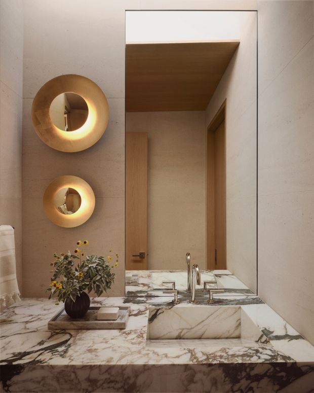 A refined bathroom vanity viewed straight on, featuring a large frameless mirror, warm beige stone wall panels, a dramatic veined marble countertop and integrated rectangular basin. Brushed brass-toned wall lights and polished metal tapware provide warm accents, with timber cabinetry and ceiling visible in the mirror reflection. A small floral arrangement sits on the vanity.