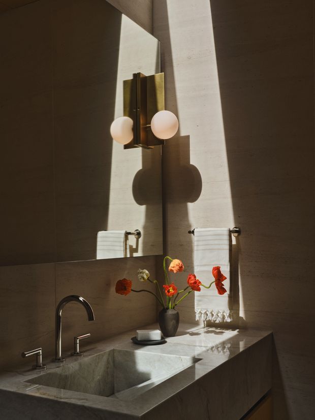 A refined, moody bathroom vanity in warm stone, featuring an integrated rectangular basin, dark curved faucet, large mirror, brass wall sconce with globe bulbs, and white hand towels. Strong sunlight creates dramatic shadows across the stone surfaces, with a small vase of red and orange flowers on the counter.