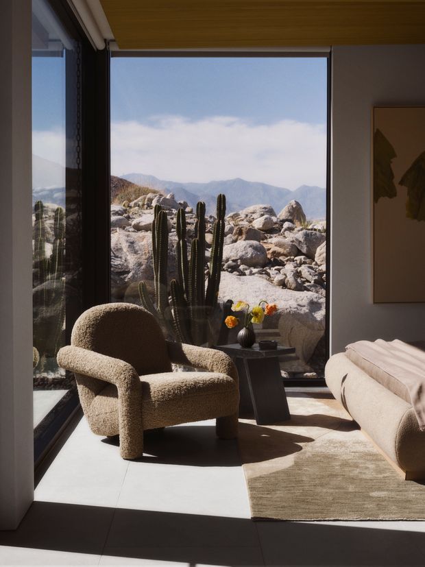 A warm, minimalist living room with a boucle lounge chair, neutral upholstered sofa, small dark side table, and textured rug. Floor-to-ceiling glazing frames a rugged desert landscape with tall cacti and distant mountains. The room has a timber-lined ceiling, dark window frames, strong natural light, and restrained neutral decor.