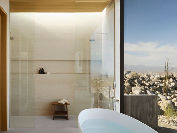 A serene contemporary bathroom with a freestanding white bathtub positioned beside full-height glazing overlooking a rocky desert-like landscape. The open walk-in shower has clear glass panels, wall-mounted shower fixtures, pale stone or large-format tile walls, a warm timber ceiling, and a small wooden stool with toiletries. The room is bright, minimal, and strongly connected to the exterior view.