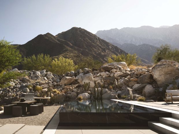 A landscaped outdoor pool terrace set against rugged desert mountains. The scene features a dark rectangular reflecting or swimming pool, pale paved decking, outdoor lounge furniture, a built-in barbecue or outdoor kitchen area, boulders, and drought-tolerant desert planting.
