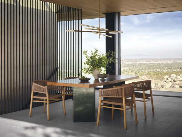 Contemporary dining area with a dark stone or metal dining table, woven timber dining chairs, and a sculptural linear pendant light. Full-height glazing opens to a broad landscape view, while vertical black screening and timber-lined ceilings add architectural texture.