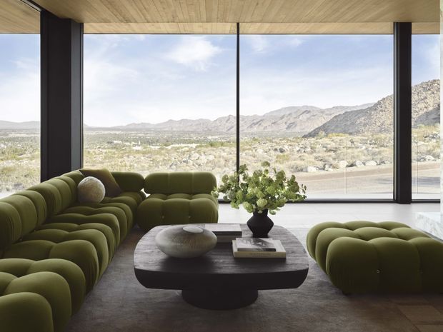 A sophisticated living room with expansive floor-to-ceiling glazing overlooking a rugged mountainous desert landscape. The room features a large sculptural olive-green modular sofa, matching rounded lounge seating, a dark sculptural coffee table, a neutral rug, and a timber-lined ceiling. Dark vertical window frames create a strong architectural rhythm, while abundant natural light gives the space a refined, organic character.