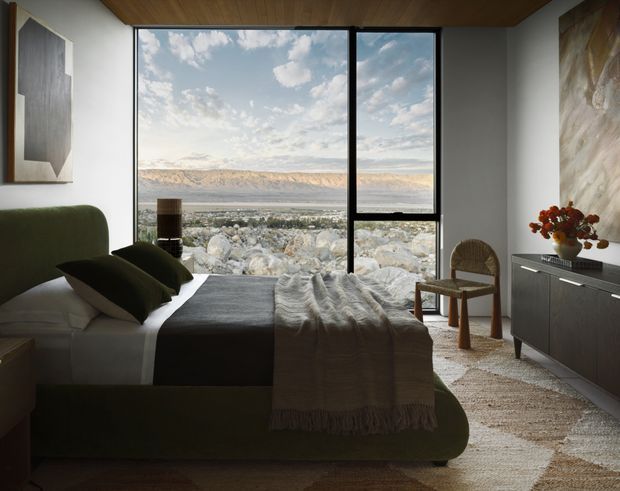 A sophisticated bedroom with a low-profile olive-green upholstered bed, dark bedding, and a textured throw. Full-height black-framed glazing provides expansive views across a rocky foreground and broad mountainous landscape. The room features a warm timber ceiling, light neutral walls, a dark built-in sideboard, woven rug, sculptural artwork, and a small timber chair.