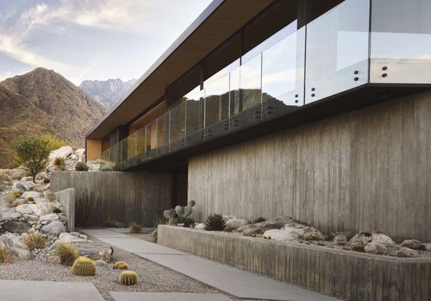 A modern, low-profile building set against rugged mountains, featuring a long cantilevered roof, extensive floor-to-ceiling glazing, frameless glass balustrades, and textured concrete walls. The landscaped foreground uses gravel, boulders, cacti, and drought-tolerant planting alongside a concrete walkway.
